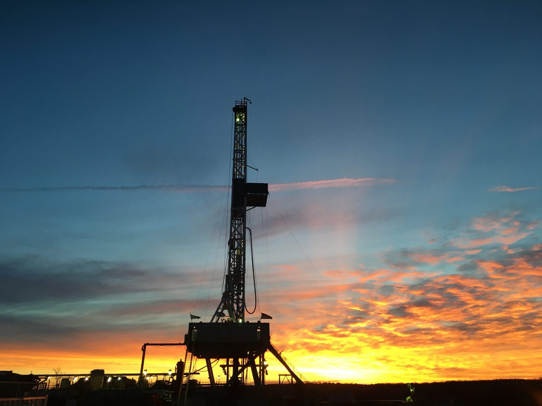 Land drilling rig against an orange sunset, rig-floor lights on.